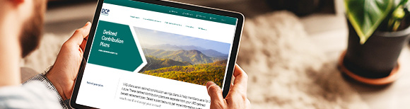 An over the shoulder view of a man seated on a couch while holding a tablet upon which the Virginia Retirement System DCP website is displayed.