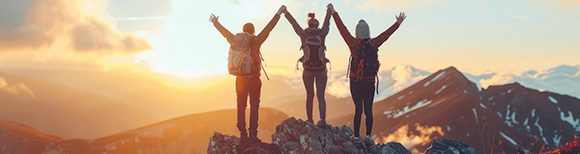 Rear view of three people standing on the summit of a mountain with their arms raised in triumph, overlooking the sunrise.