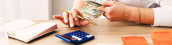 a pair of hands holding cash and a pen. A calendar book, calculator and other papers are in front of them on a tabletop.