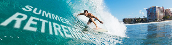 A man rides a surf board across a large sea wave with text on it saying summer retirements. In the background there is a sandy beach with large hotel buildings along the back. The weather is sunny and clear.