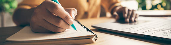 Decorative. A hand writes with a pen in a notebook resting on a desk. The other hand rests on the keyboard of a laptop computer on the desk. The person's torso is in the background.