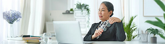 Decorative. An older woman sitting at her desk in her office. Several potted plants are on surfaces around her.