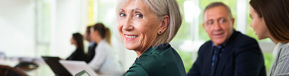 Decorative. An older woman turned to look over her shoulder and smile at the viewer while seated at a conference room table . Others in the background are seated at the same table.