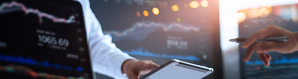 Three computer desktop monitors show upward investment trend lines. Between them, one person's hand at the end of a long sleeve touches a smaller computer tablet. On the opposite side of the frame, another hand holds a writing pen. Illustrative only.