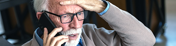 An older man with white hair and beard wears eyeglasses while holding a smartphone to his right ear. He has a concerned expression on his face and holds his left hand on the top of his head in distress. Illustrative only.