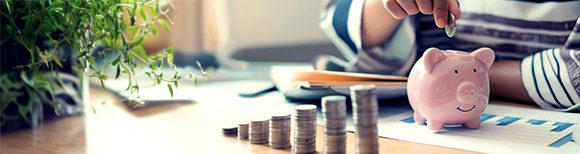 A person places a coin into a ceramic piggy bank that rests on top of financial papers on a wooden tabletop. In front of the piggy bank are ascending stacks of coins. On the left side of the frame is a large house plant. Illustrative only.