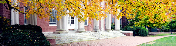 Low stairs, a stone porch with large stone columns with windows beyond them and a brick walkway with lawn are framed by low-hanging tree branches with autumn leaves at the University of Mary Washington in Fredericksburg, Virginia. Illustrative only.