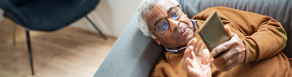 An older man wearing glasses and a sweatshirt lies on a couch holding a cellphone above himself with both hands.