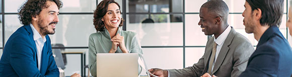 Office workers around a conference room smile at their colleague seated behind her laptop.