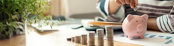 Decorative. A young person sits at a table placing coins into a ceramic piggy bank. On the table in front of them are financial papers and ascending stacks of coins. On the table to the left is a large, green houseplant.