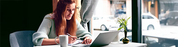 Decorative. A young woman sitting at a table near a large window in a cafe. She is looking at her laptop computer screen and writing in a small notebook. Outside the window are cars parked near the curb and buildings on a city street.