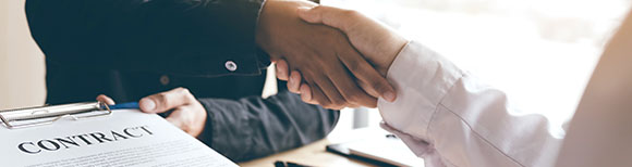 Decorative. Two people shaking hands in agreement across a desk while one holds a contract on a clipboard in their opposite hand. Pens rest upon the desktop. There is a window in the background..