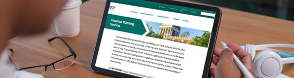 Decorative. An over the shoulder view of a man seated at a small table holding a computer tablet. On its screen is displayed  the DCP Financial Planning Services website. On the table are a single use coffee cup with a lid, a pair of eyeglasses and a pair of over the ear headphones.