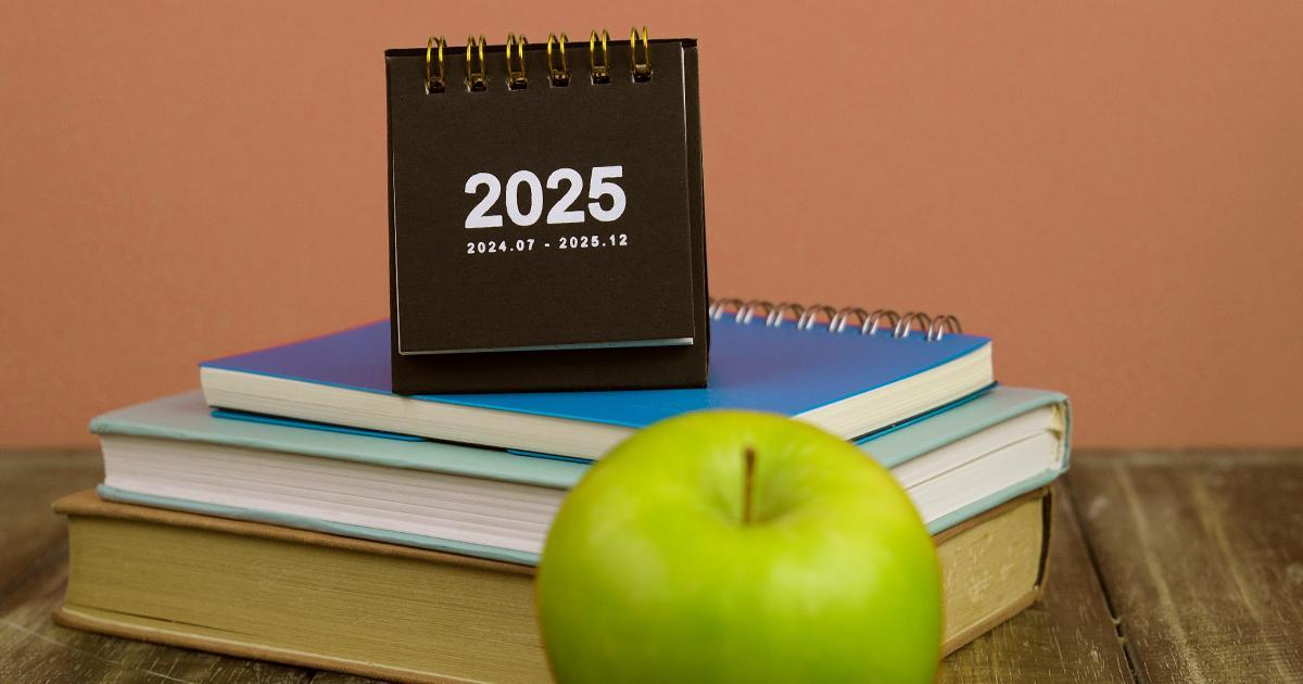 Decorative. A tabletop with a small 2025 calendar upright on top of a stack of books. In the foreground is a large apple.