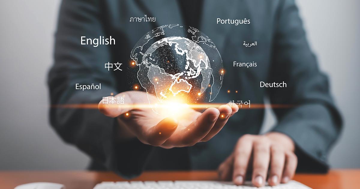 Illustrative. A male torso in professional dress stands in the background with his left hand on a tabletop. He opens his right hand, palm upward toward the viewer. Superimposed over his open hand is a graphic image of the land masses of the Earth and a glowing light. Around the Earth graphic are listed names of languages.