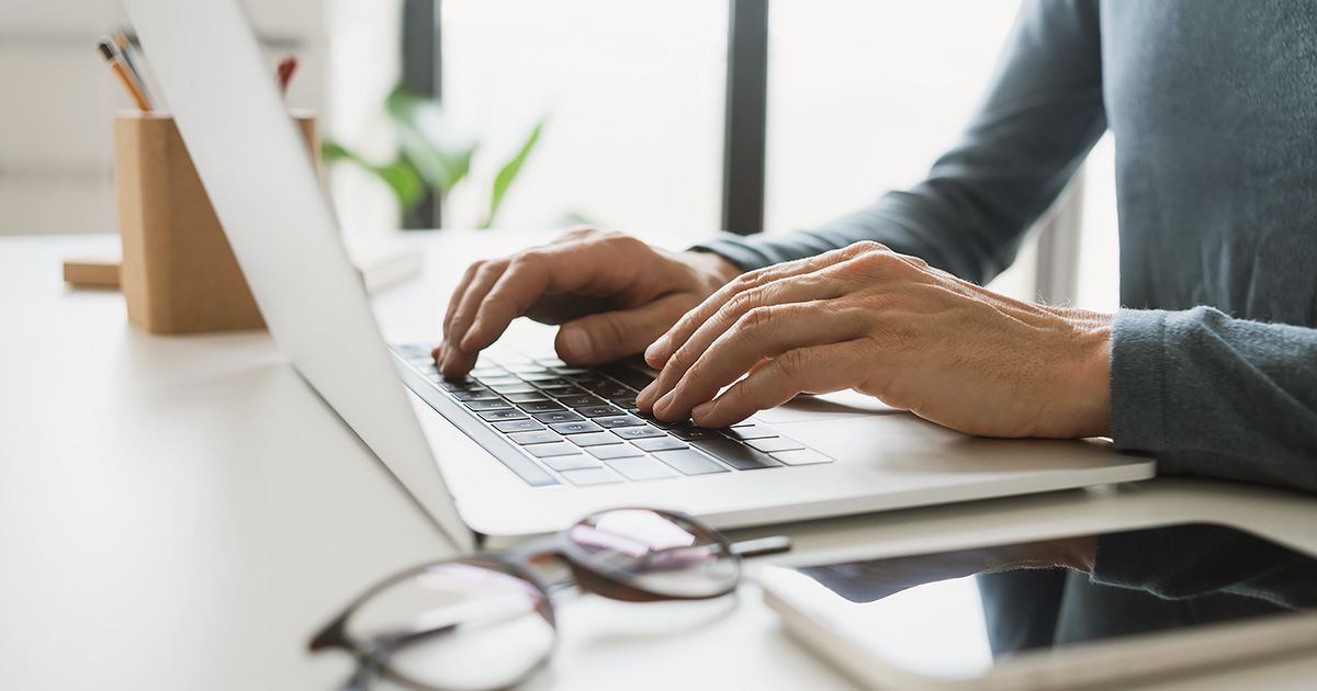 Decorative. A man's torso, arms and hands are on the right. He types on a laptop computer on a tabletop. In the foreground are a pair of folded eyeglasses and a smart phone. In the background are a wooden rectangular pencil holder with pencils, a plant and a several large windows.