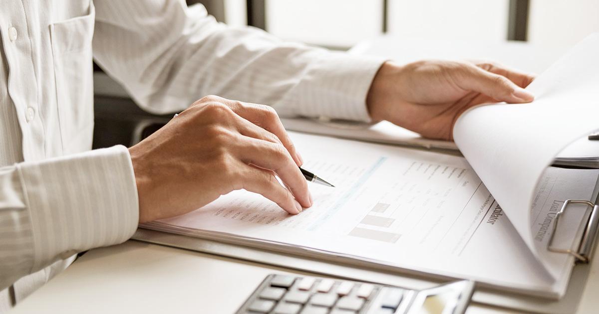 Decorative. On the left, a man's torso, arms and hands rest on a tabletop with a clipboard. Attached to the clipboard is a thick stack of financial charts. In his nearest hand, he holds a writing pen between his fingers. In the foreground is a partial view of a calculator. In the background are several large windows.