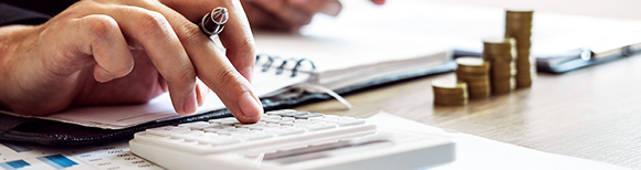 Closeup view of a hand typing on a calculator on a desk while papers and stacks of coins lie nearby.