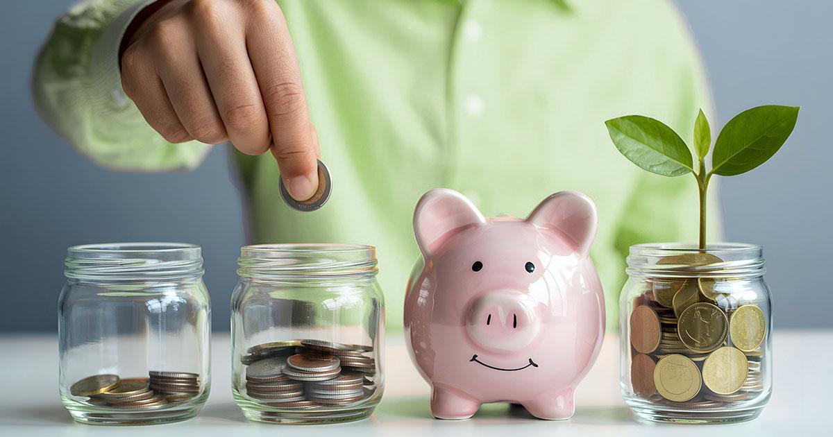 Decorative. A man seated at a table, wearing a dress shirt with long sleeves, places coins into the second small jar in a line of three small jars and a small ceramic piggy bank. A seedling grows from the last jar which is full of coins.