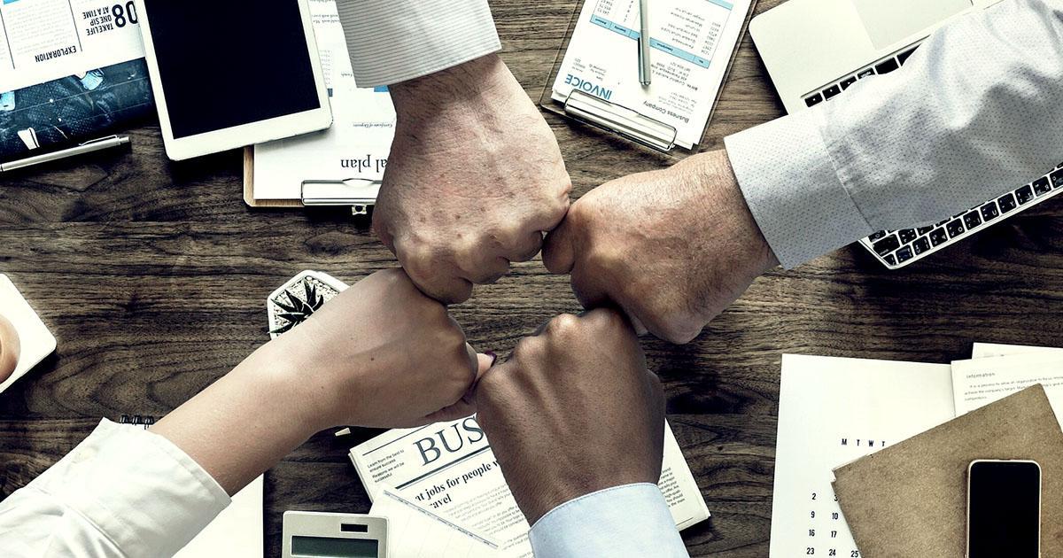 Decorative. View from above of the fists of four office workers bumping together in a gesture of celebration or solidarity. On the table surface beneath them are notebooks, clipboards, a computer tablet, keyboard and a calculator.