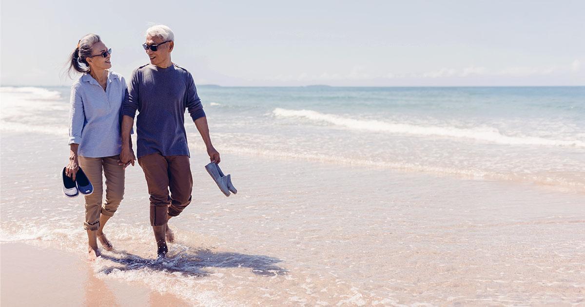 Decorative. A retired couple walk along a sunny beach barefoot while carrying their shoes.