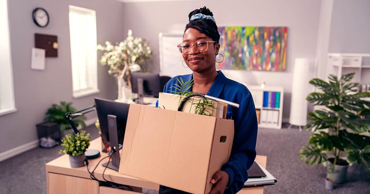 Decorative. A smiling woman stands in a room with office desks. She is carrying a cardboard box full of desk items.