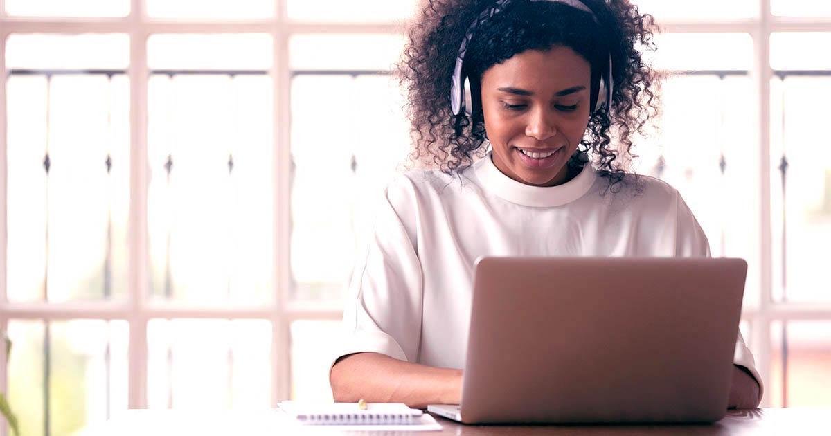 Decorative. A young woman seated at a desks wears over-the-ear headphones and smiles down at her laptop computer. Behind her is a wall of bright windows.