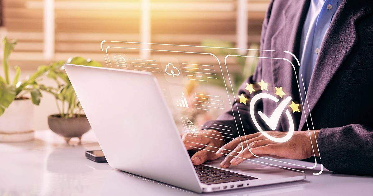 Decorative. A man's professionally dressed torso and hands on the far right are seated at a desk with potted houseplants on the far left. He types on his laptop computer. SUperimposed between him and the screen is a graphic of a page of text with icons and a larger icon of a check mark in a circle with five starts in an arc around the top of the circle. This indicates success.