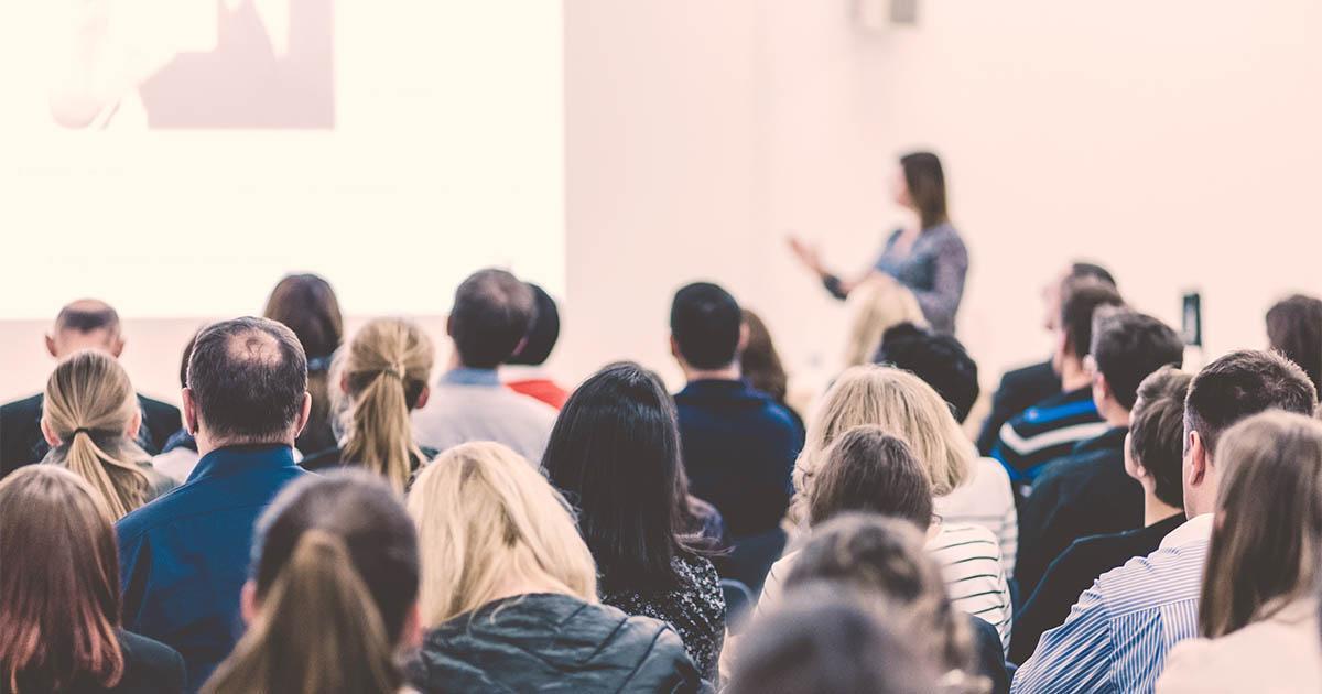Decorative. Rear view of a large room with several rows of people in chairs facing a woman speaking to them and gesturing to a large financial chart projected onto a screen.