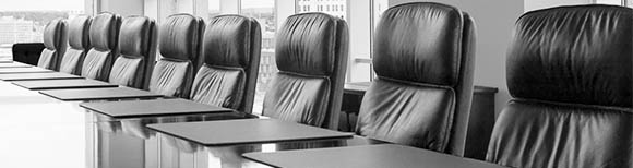 Decorative. A row of matching executive chairs line one side of a long conference room table. Windows and a city skyline are in the background.