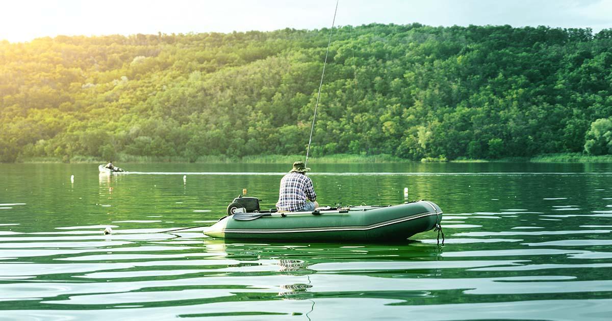 Decorative. Rear view of a man who is fishing on a boat in the middle of a small lake. Trees and mountains are in the background.