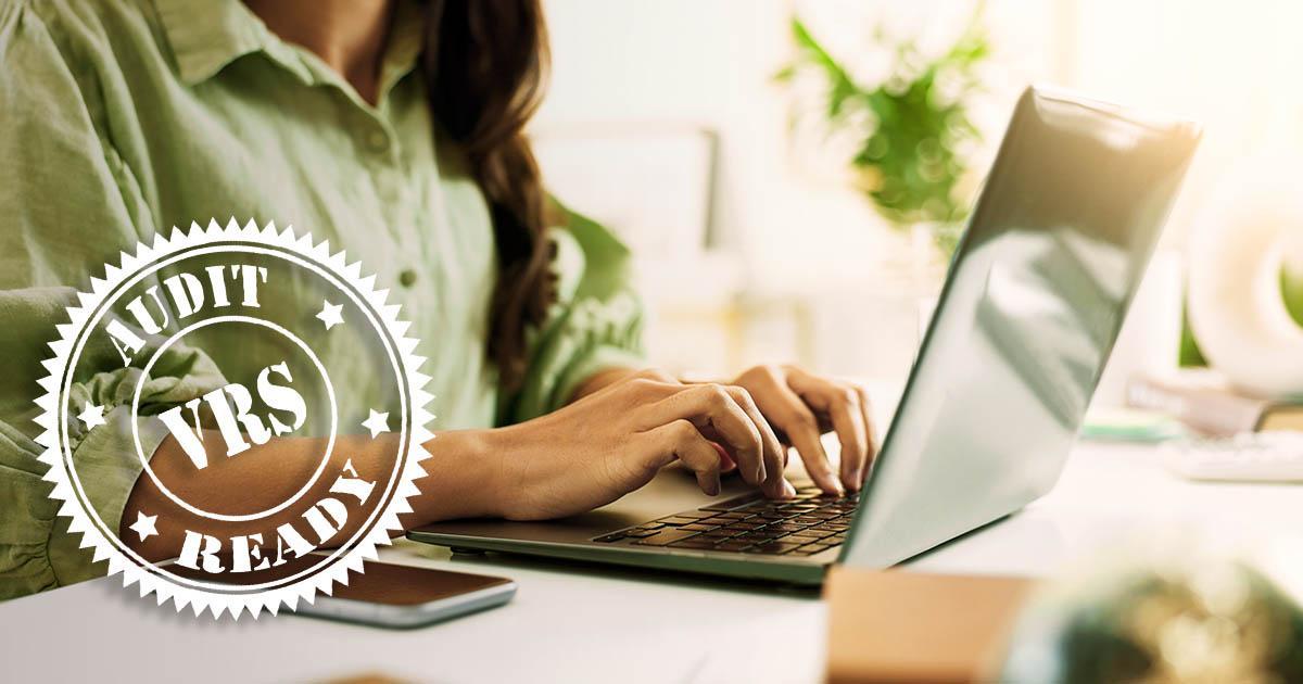 Decorative. A photo of a woman's hands and torso seated at a desk, working on her laptop with her cell phone nearby. Potted house plants are in the background. In the lower left corner is a round graphic which reads VRS Audit Ready overtop of the photo.