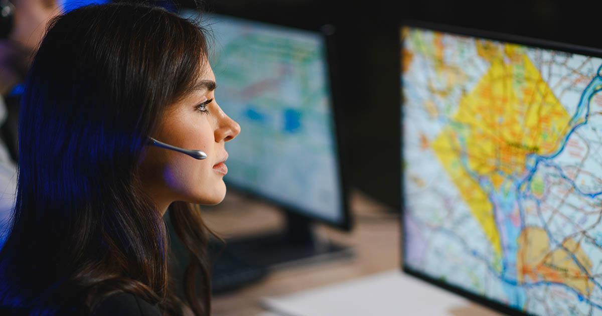 Decorative. A young woman wearing a headset with microphone looks at her computer monitor which shows a map of a city.