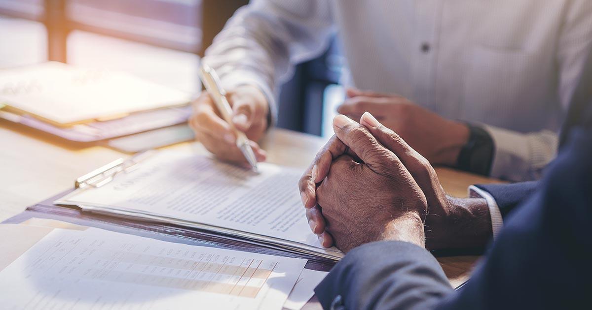Decorative. A closeup view of two torsos and pairs of hands going over contract papers on the table at which they are seated.