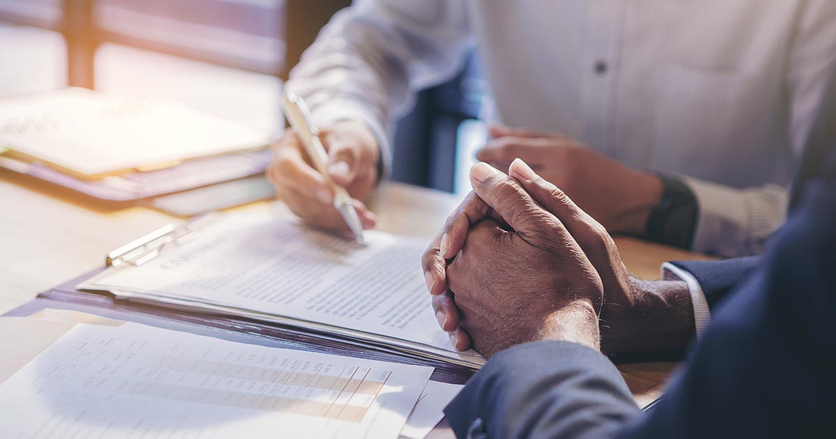Decorative. A closeup view of two torsos and pairs of hands going over contract papers on the table at which they are seated.