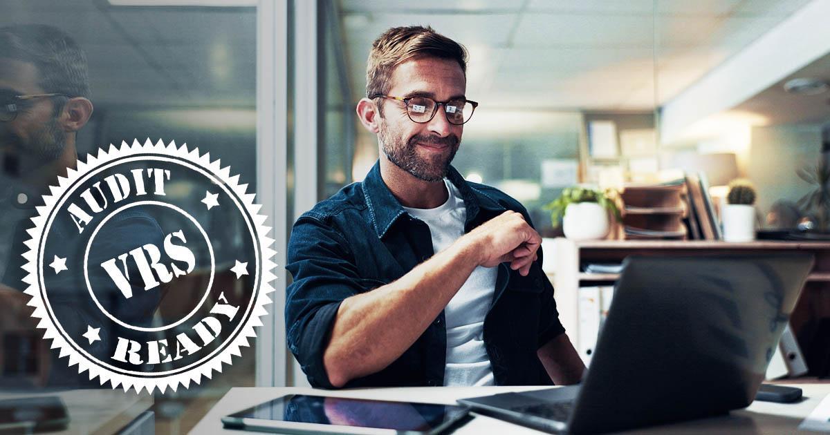 Decorative. A man in casual dress its at a desk with a laptop computer and computer tablet before him. On the far left is superimposed a round graphic that reads VRS audit ready.