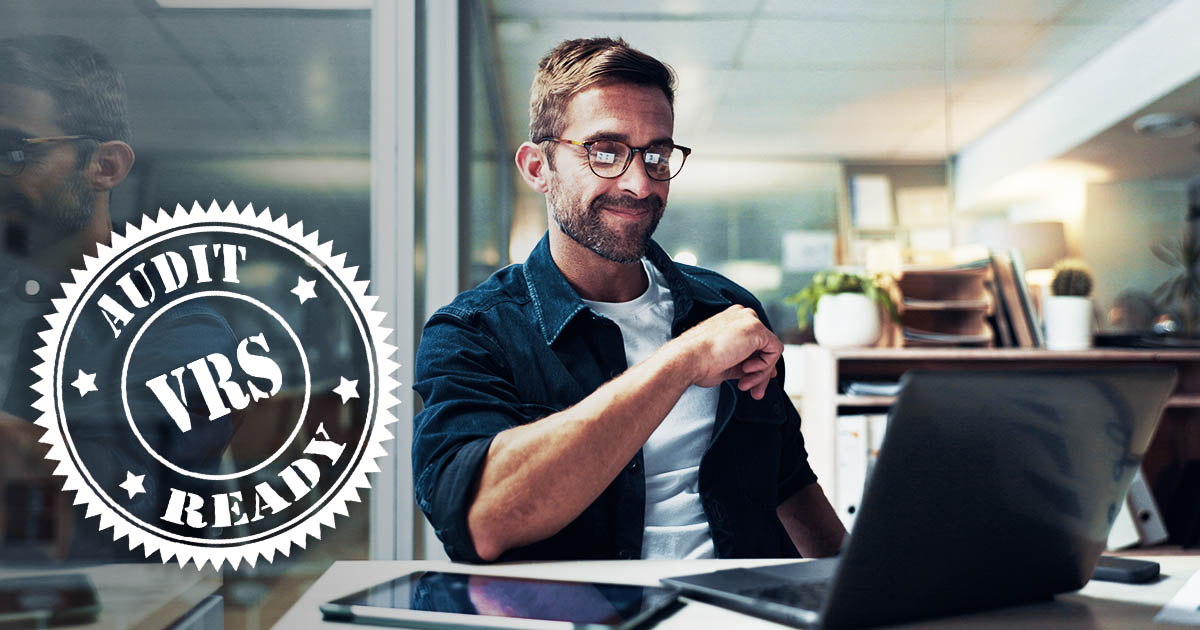 Decorative. A man in casual dress its at a desk with a laptop computer and computer tablet before him. On the far left is superimposed a round graphic that reads VRS audit ready.