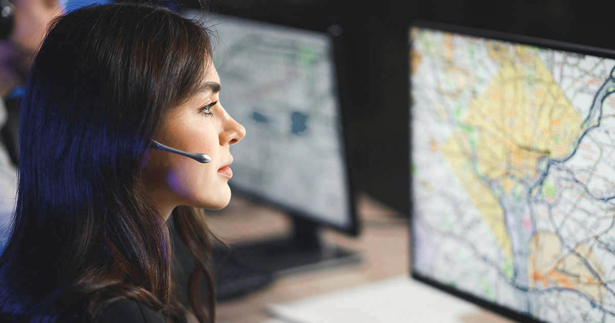 Decorative. A profile view of a woman wearing a headset with microphone is on the far left. She is looking a large computer monitor on the right showing a layout of city streets.