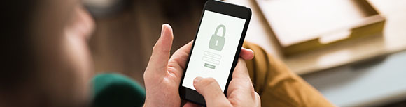 Decorative. Over-the-shoulder view of a person holding a smartphone in both hands. On the phone screen is a graphic of a padlock and a password field.