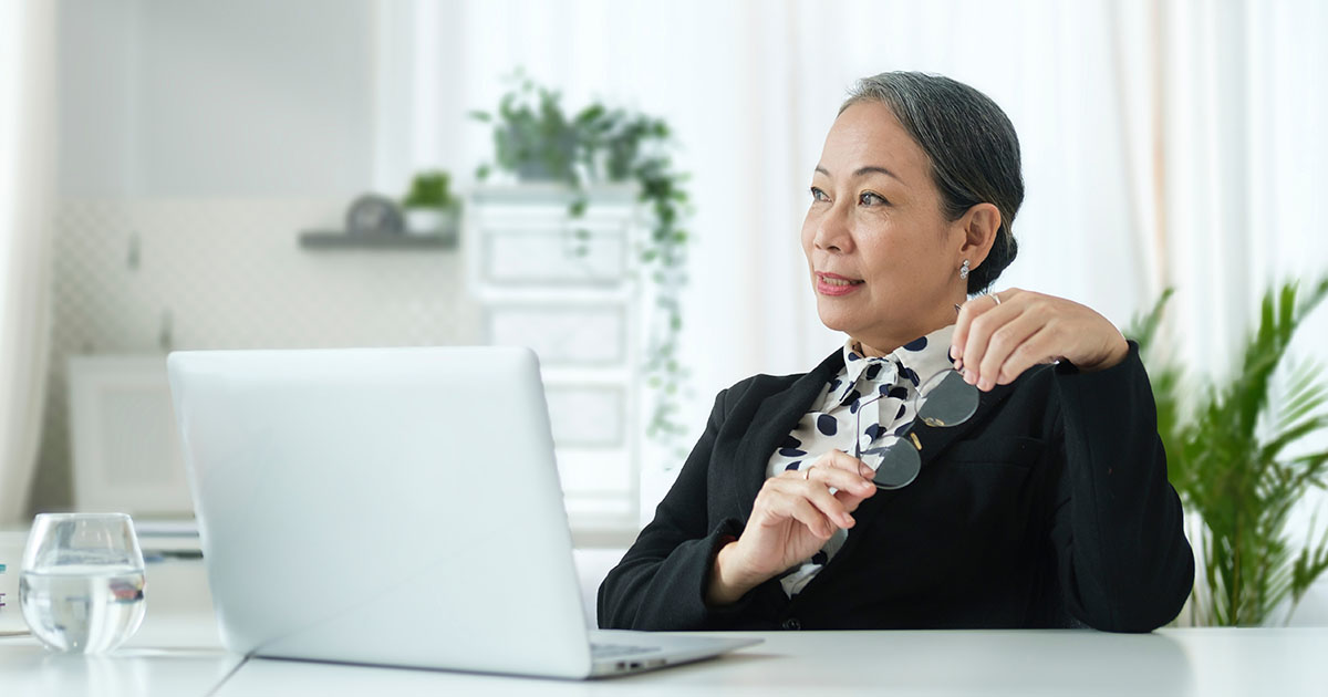 Decorative. An older woman sitting at her desk in her office. Several potted plants are on surfaces around her.