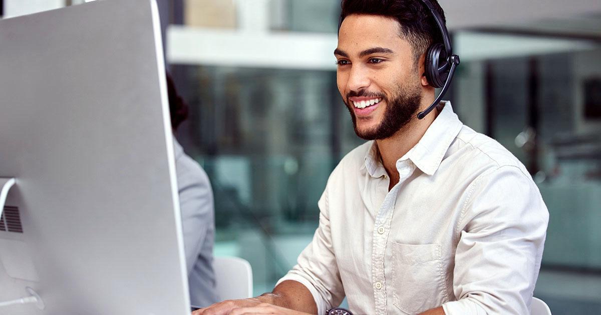 Decorative. A young man in an office smiles at his computer screen while wearing headphones with an attached microphone.