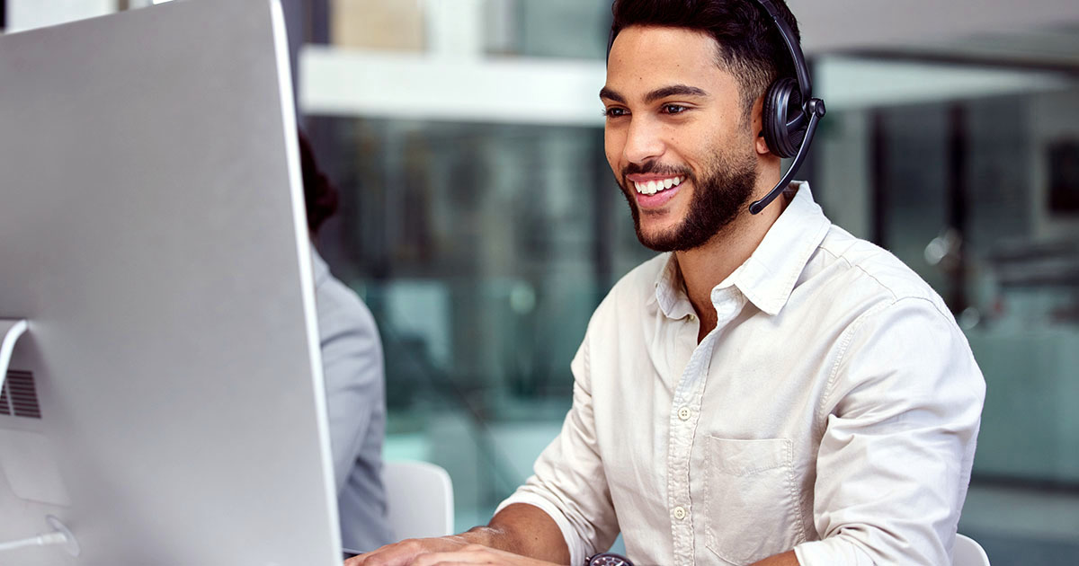 Decorative. A young man in an office smiles at his computer screen while wearing headphones with an attached microphone.