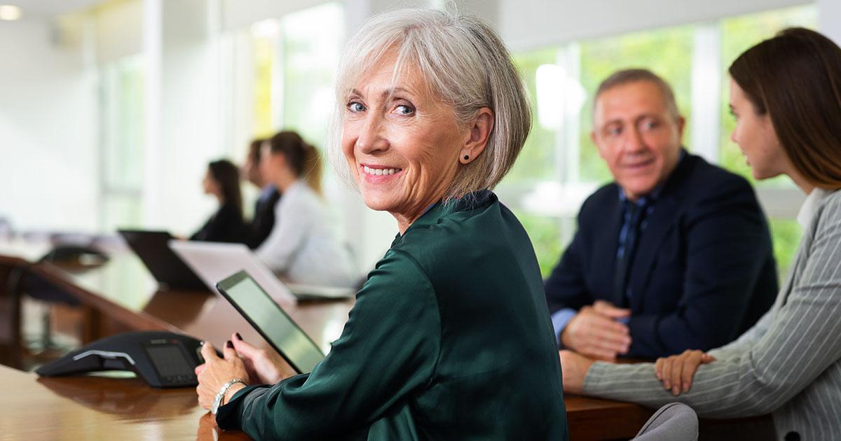 Decorative. An older woman turned to look over her shoulder and smile at the viewer while seated at a conference room table . Others in the background are seated at the same table.