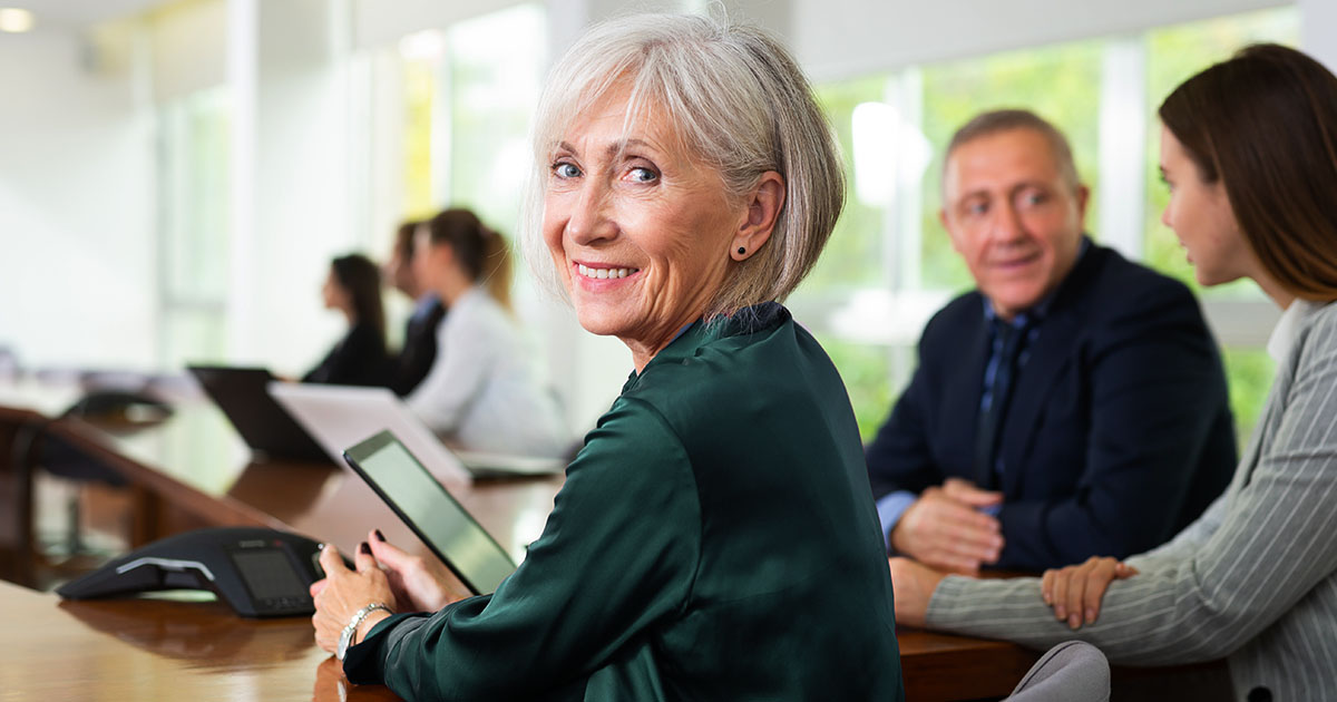 Decorative. An older woman turned to look over her shoulder and smile at the viewer while seated at a conference room table . Others in the background are seated at the same table.