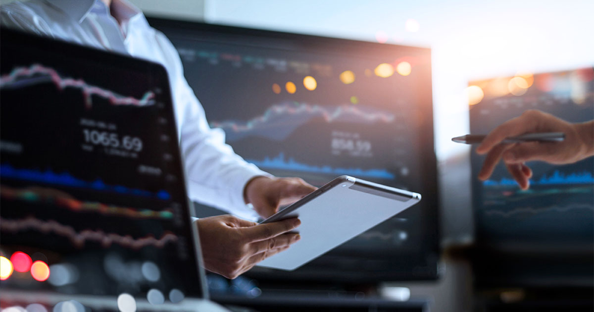 Three computer desktop monitors show upward investment trend lines. Between them, one person