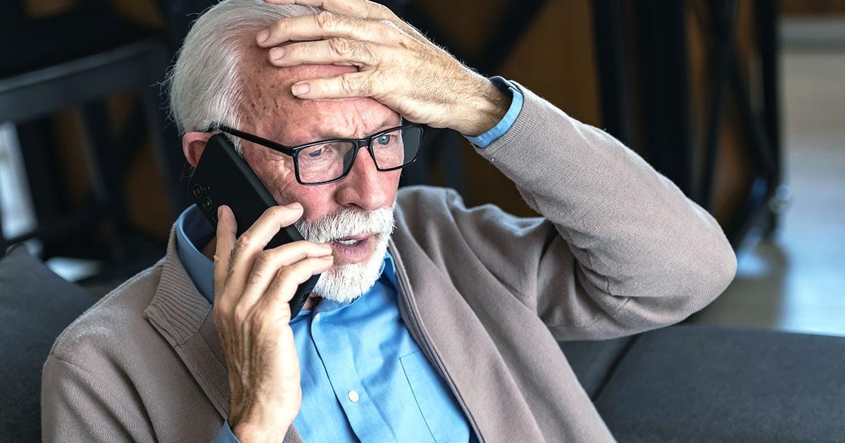 An older man with white hair and beard wears eyeglasses while holding a smartphone to his right ear. He has a concerned expression on his face and holds his left hand on the top of his head in distress. Illustrative only.