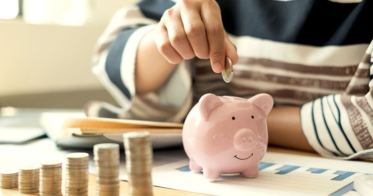 A person places a coin into a ceramic piggy bank that rests on top of financial papers on a wooden tabletop. In front of the piggy bank are ascending stacks of coins. On the left side of the frame is a large house plant. Illustrative only.