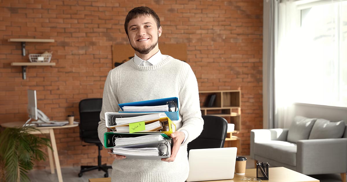 Decorative. A smiling young man wearing a long sleeved sweater stands in a casual office with several desks. He carries several large binders full of papers.