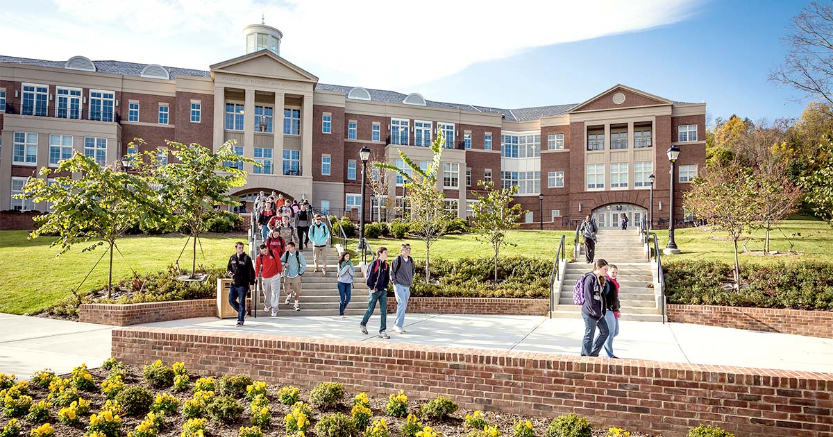 Decorative. Outdoor view of the facade of the College of Business and Economics at Radford University in Radford, Virginia. In the foreground, a group of students exit the building down stairs and sidewalks surrounded by manicured landscaping.