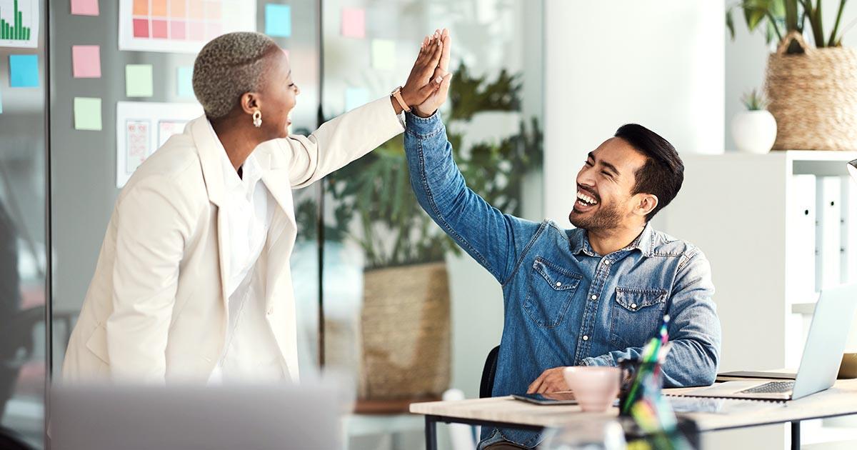 Decorative. In a modern office with glass walls, a standing woman on the left high-fives a smiling man seated at a desk on the right.